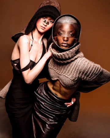 Two women pose in futuristic high fashion: one in a helmet and turtleneck, the other in a dress and hat, against a warm-toned backdrop.