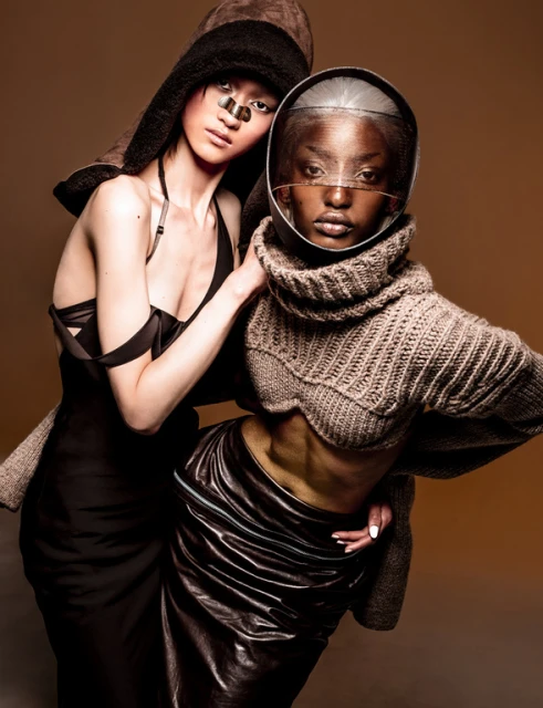 Two women pose in a studio, one wearing a brown hooded hat, the other a clear visor helmet and knit top, against a tan backdrop.