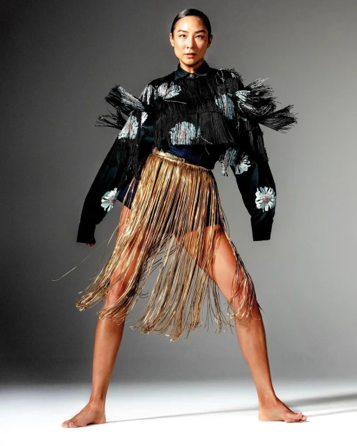 A full-length studio portrait of an Asian woman modeling an unusual fringed black and gold outfit against a neutral gray background.