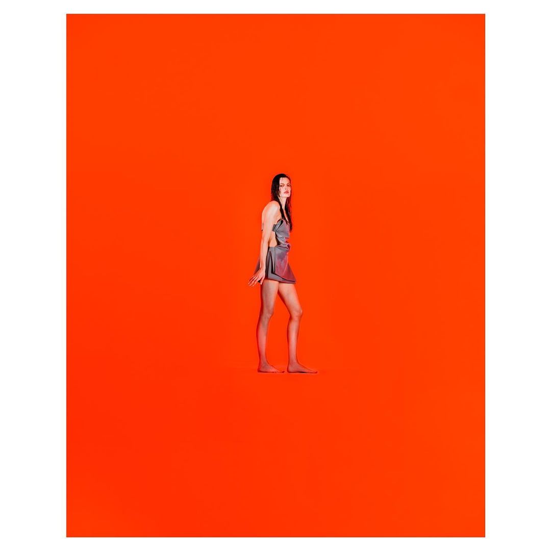 A woman wearing a metallic mini dress stands barefoot against a vibrant orange studio background.