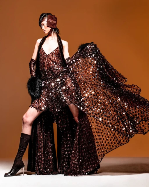 A stylish Asian female model poses in a sparkly brown dress, matching gloves, and boots against an orange background in a studio setting.