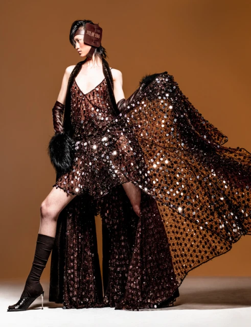 A stylish woman models a brown sequin dress with a matching cape and boots in a studio setting. Fashion, glamour, and elegance are highlighted.
