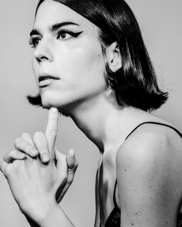 Black and white portrait of adult female with chin resting on finger, dark hair, and eye makeup, set against a neutral background.