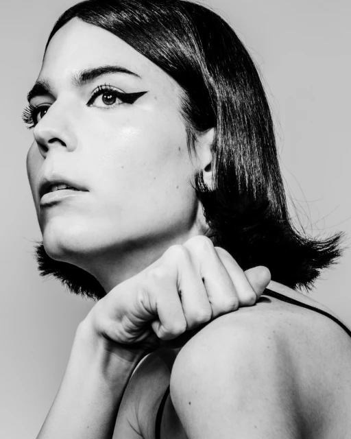 Striking black and white portrait of a woman with bob haircut and bold eyeliner, hand raised near shoulder, against a neutral background.