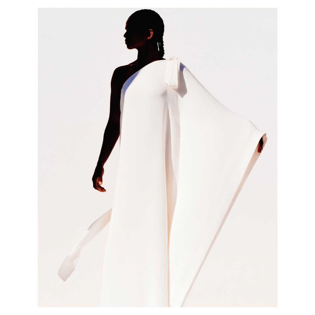 Elegant portrait of a woman wearing a flowing white gown against a white background, captured in soft studio lighting with shadow.