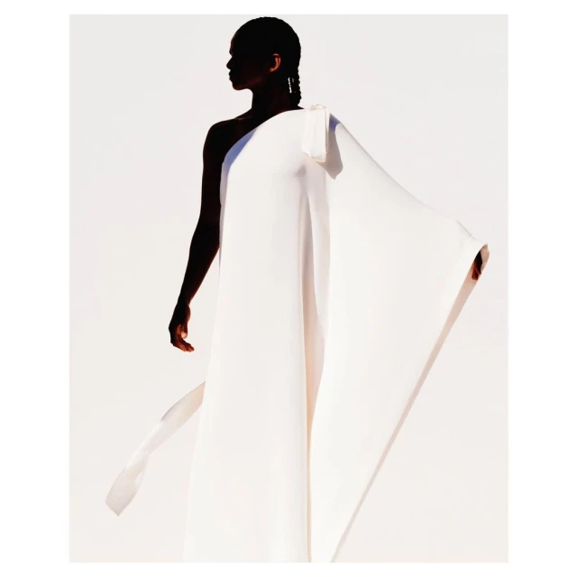 Elegant portrait of a woman wearing a flowing white gown against a white background, captured in soft studio lighting with shadow.