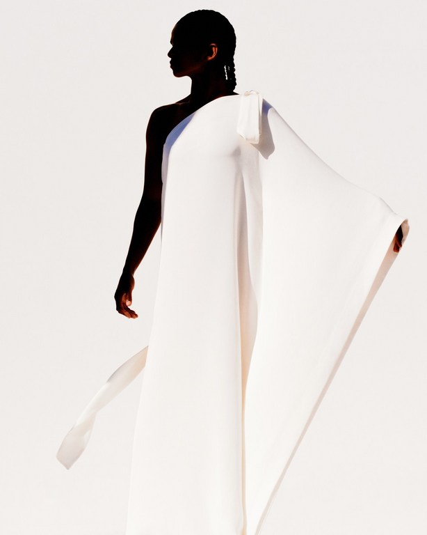 Model in flowing white one-shoulder gown standing against a plain bright white studio background.