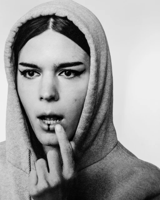 Black and white portrait of a person with dark hair and striking winged eyeliner, wearing a hooded sweatshirt and touching their lip.