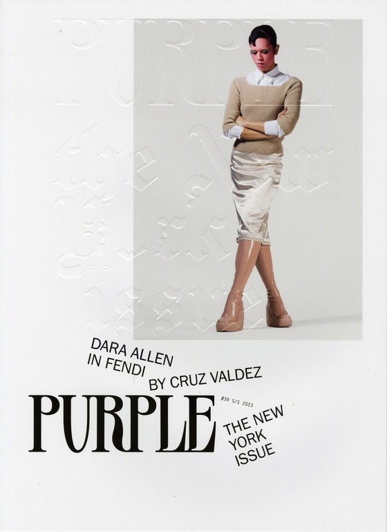 Fashion editorial shot of a model in a tan sweater and skirt, wearing tan boots, posing indoors against a textured white backdrop.