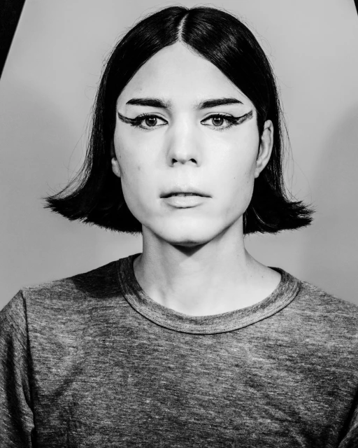 Monochrome portrait of a person with short black hair and dramatic black eyeliner wearing a grey long-sleeved shirt against a grey background.