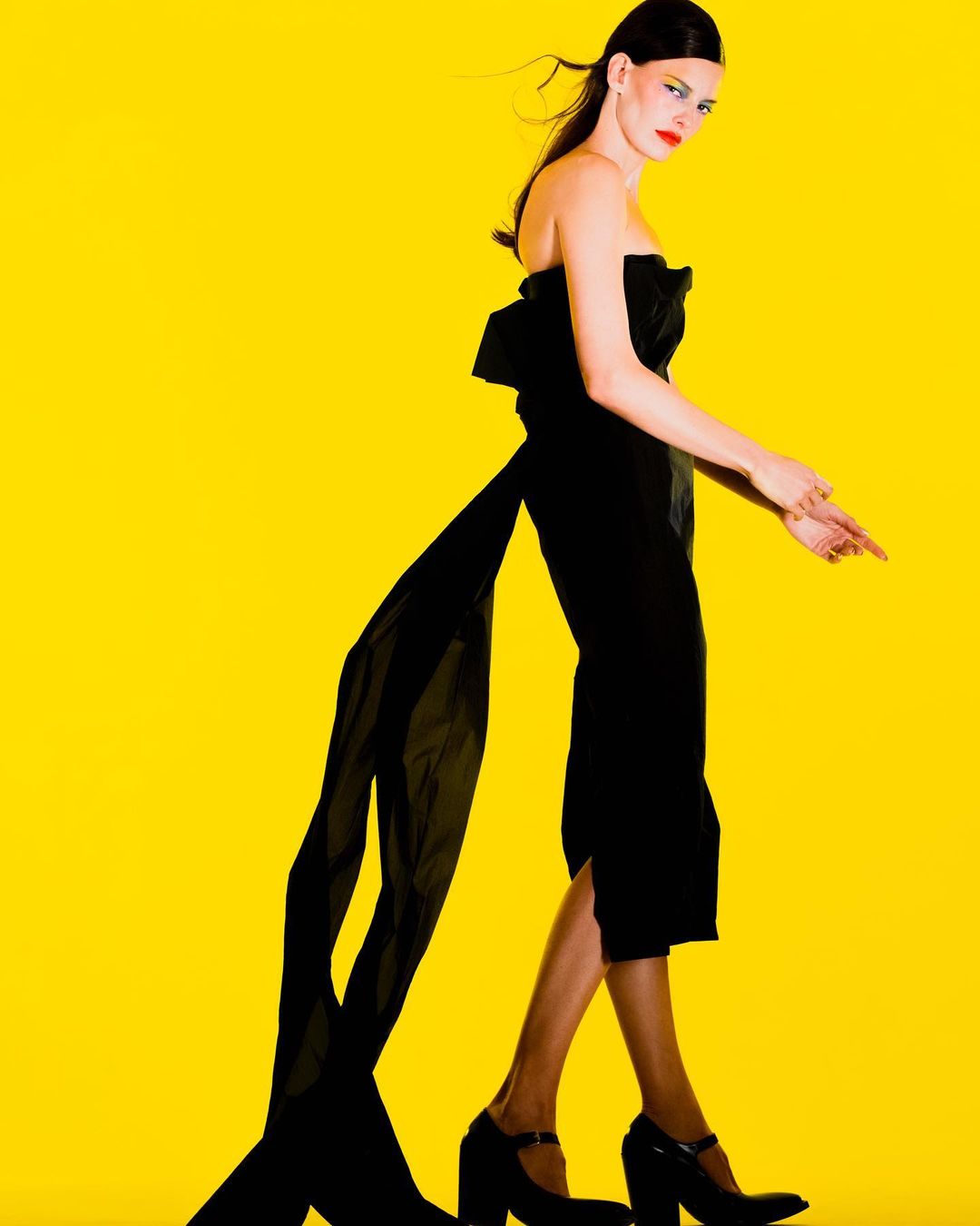 A woman in a black dress with a long train poses in a studio with a vibrant yellow background, showcasing fashion and style.