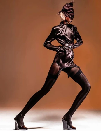 Model in dark satin dress, thigh-high stockings, and headpiece photographed by Cruz Valdez against warm gradient background.