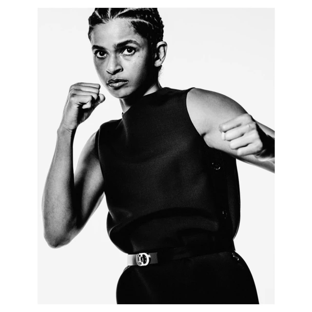 Monochromatic portrait of a woman in a sleeveless black top and stylish belt, posed with fists up in a boxing stance against a white backdrop.