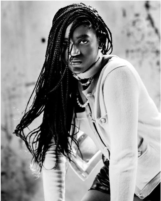 Black and white portrait of a stylish young woman with braided hair, wearing a choker and jacket, striking a fashion pose indoors.