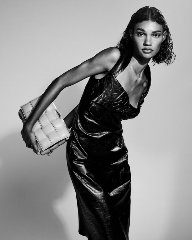 Fashion shot of a woman with curly hair, wearing a black shiny dress and carrying a woven leather bag against a grey backdrop.