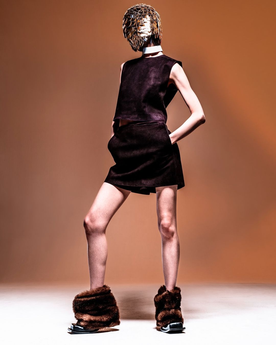A person in a dark brown dress, decorative mask, and furry shoes stands in a studio with an orange background.