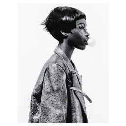 Black woman with short blunt-cut hair blowing a bubble gum bubble, wearing a textured leather jacket.