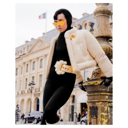 Model in cream cropped puffer jacket and yellow sunglasses leaning against an ornate stone lamppost in Paris by Cruz Valdez.