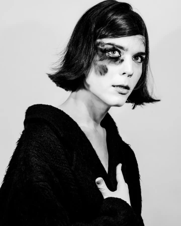 Monochrome portrait of a person with dark bob hair and dark makeup around one eye wearing a textured black top by Cruz Valdez.