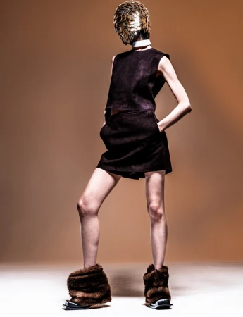 Model wearing a dark sleeveless top, dark shorts, furry boots, and a reflective golden mask against a warm backdrop