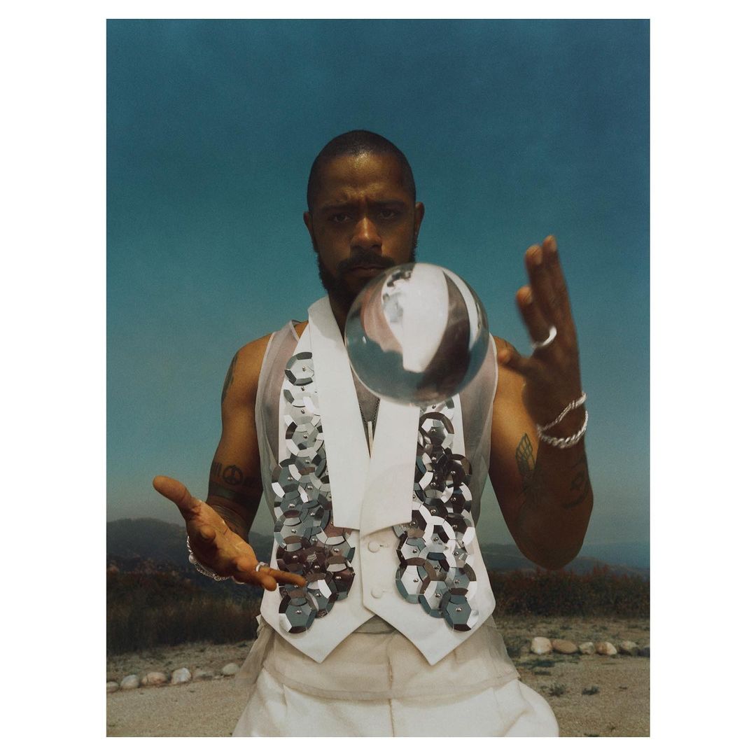Man in ornate white vest balances silver orb outdoors against a turquoise sky. He wears rings and bracelets, and his arms are outstretched.