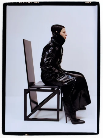 Model seated on a tall black chair wearing a glossy black trench coat and heeled boots against a white studio background.
