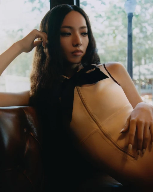 Portrait of a woman with long brown hair reclining on a leather sofa with a neutral gaze and wearing a neutral-toned dress.