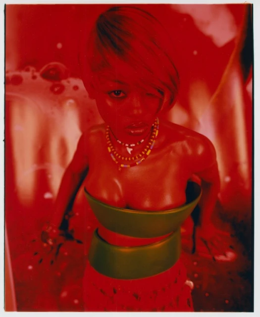 Digitally rendered woman with short blonde hair, wearing a two-piece outfit with beaded necklaces, against a red and orange background.