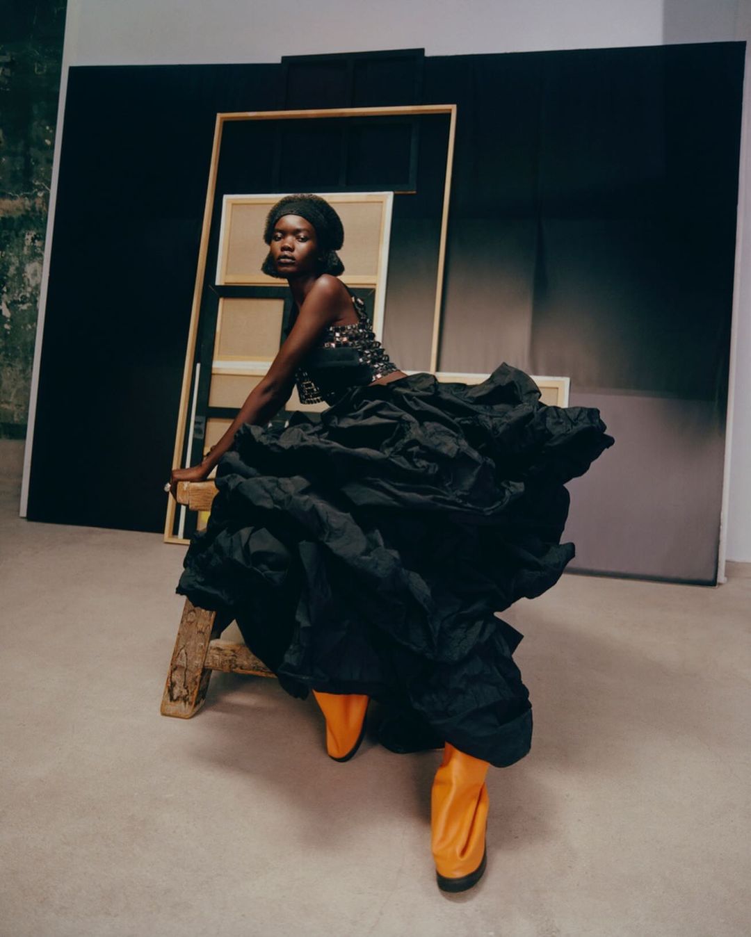 Fashion shot of a Black woman in a black dress and orange boots, sitting on a stool in front of a canvas and a dark background.