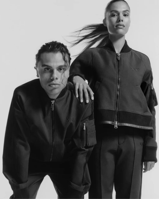 Two fashion models in matching black bomber jackets and pants pose in a studio with soft lighting, showcasing modern urban style.