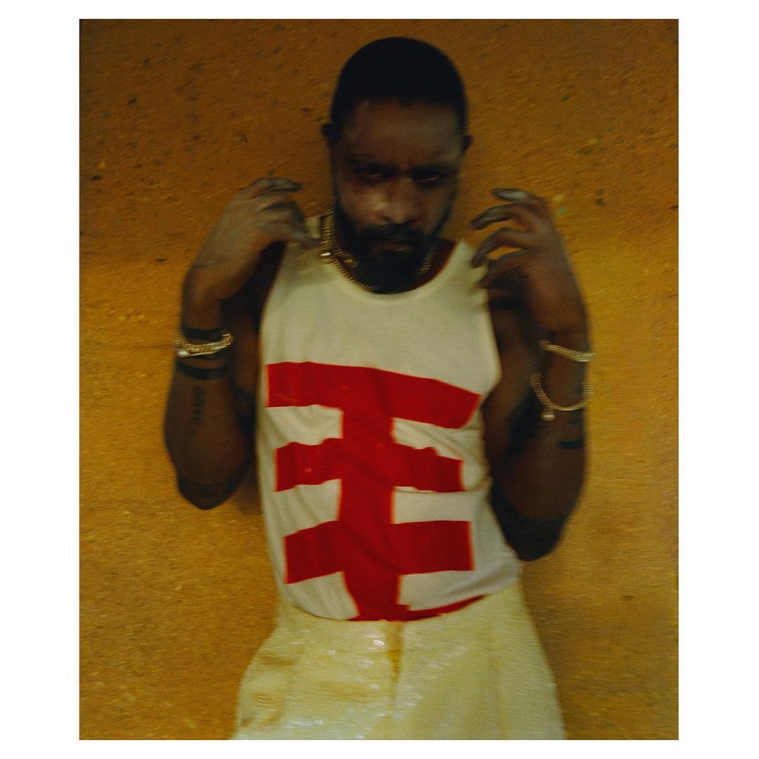 Portrait of a Black male model wearing a white tank top with a red graphic design, gold jewellery, and iridescent pants posing indoors.