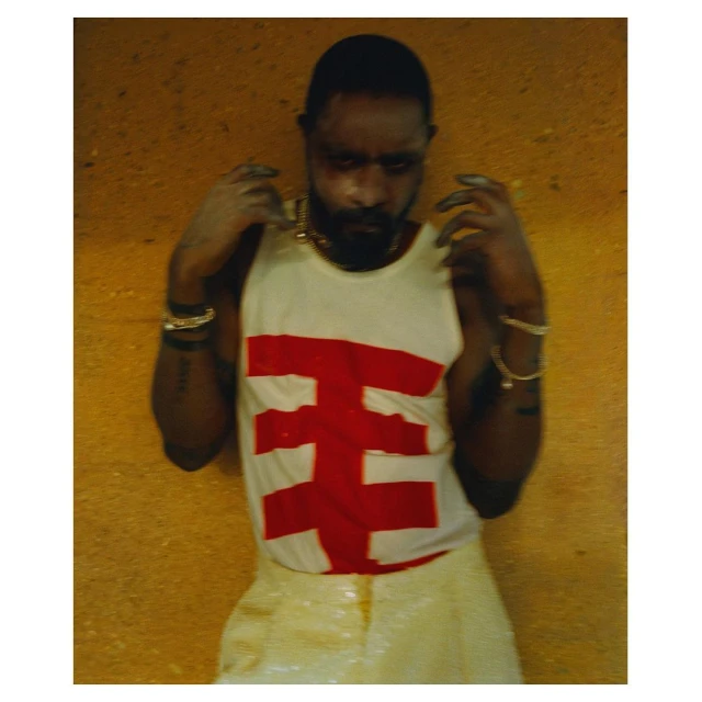 Portrait of a Black male model wearing a white tank top with a red graphic design, gold jewellery, and iridescent pants posing indoors.