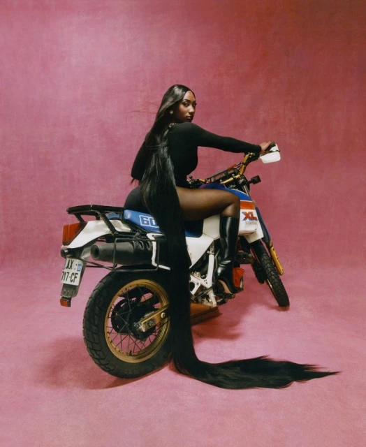 A Black woman in a bodysuit poses on a vintage motorcycle in a studio with a pink backdrop, showcasing long hair.