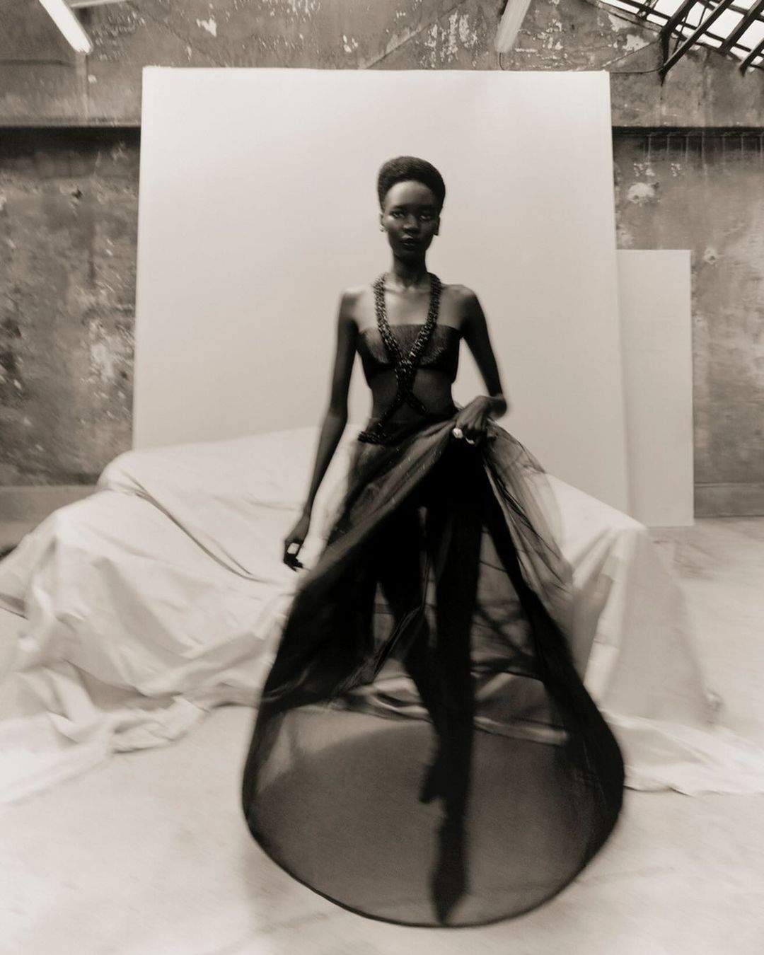 A striking black-and-white fashion shot of a Black woman in a layered black gown posing in front of a white backdrop in a spacious studio.
