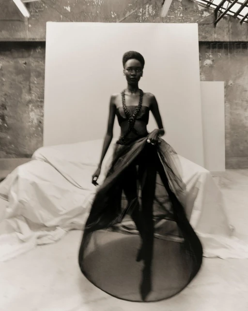A striking black-and-white fashion shot of a Black woman in a layered black gown posing in front of a white backdrop in a spacious studio.