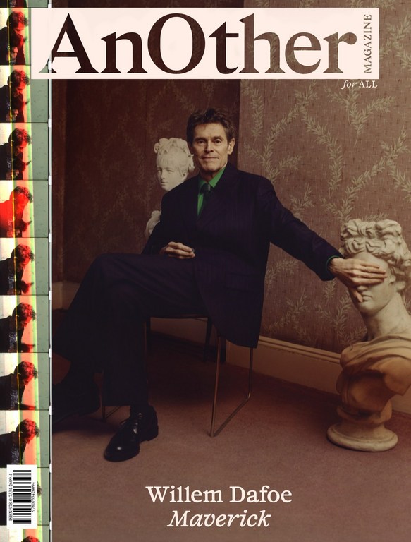 Willem Dafoe on the cover of AnOther Magazine. Actor is seated next to marble bust, wearing dark suit with green shirt.