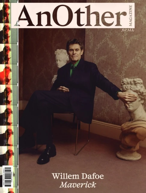 Willem Dafoe on the cover of AnOther Magazine. Actor is seated next to marble bust, wearing dark suit with green shirt.