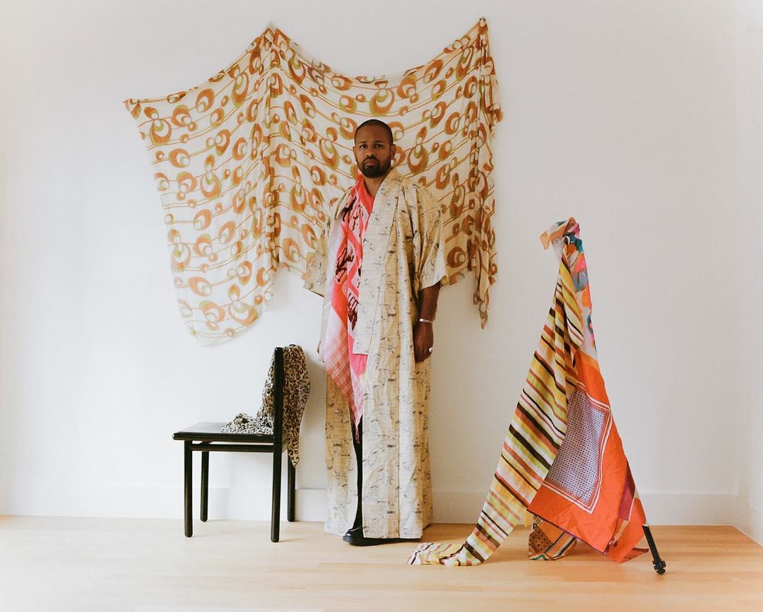 Adult man posing in a kimono in a white-walled studio with colorful draped textiles, plus black chair against a plain backdrop.
