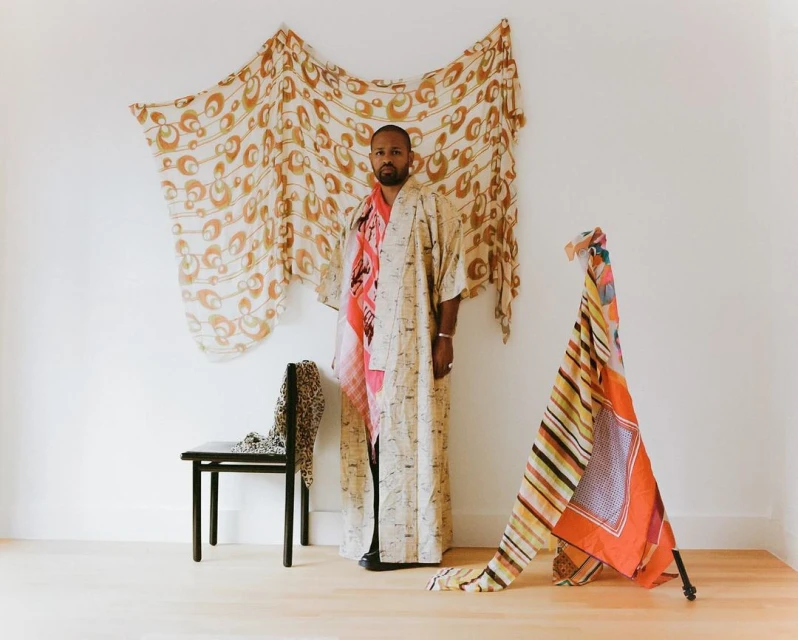 Adult man posing in a kimono in a white-walled studio with colorful draped textiles, plus black chair against a plain backdrop.