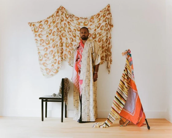 Adult man posing in a kimono in a white-walled studio with colorful draped textiles, plus black chair against a plain backdrop.