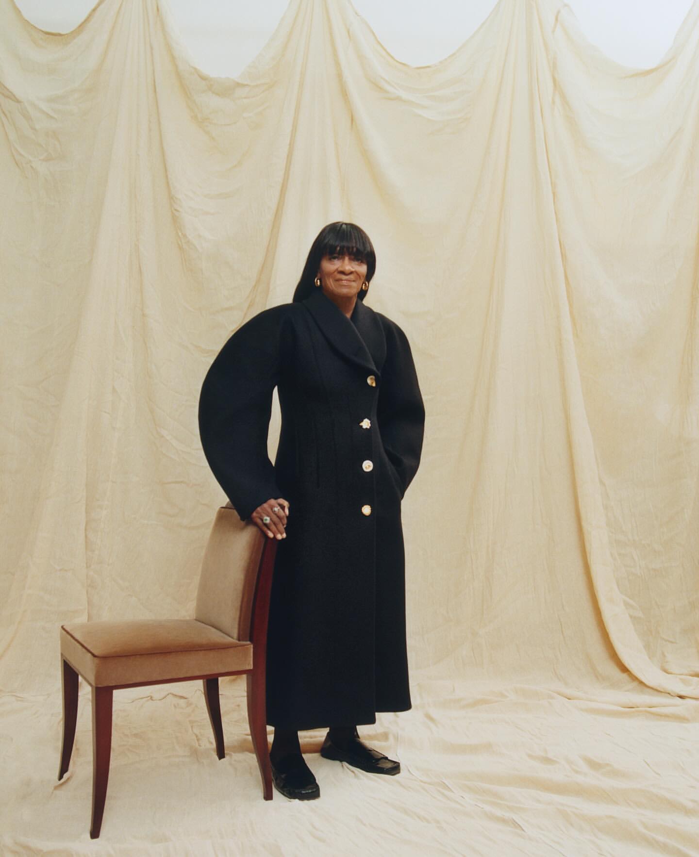 Mature African-American woman posing indoors with a black coat and a chair, standing against a draped beige backdrop, looking at the camera.