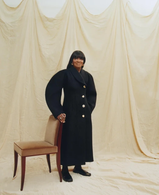 Mature African-American woman posing indoors with a black coat and a chair, standing against a draped beige backdrop, looking at the camera.