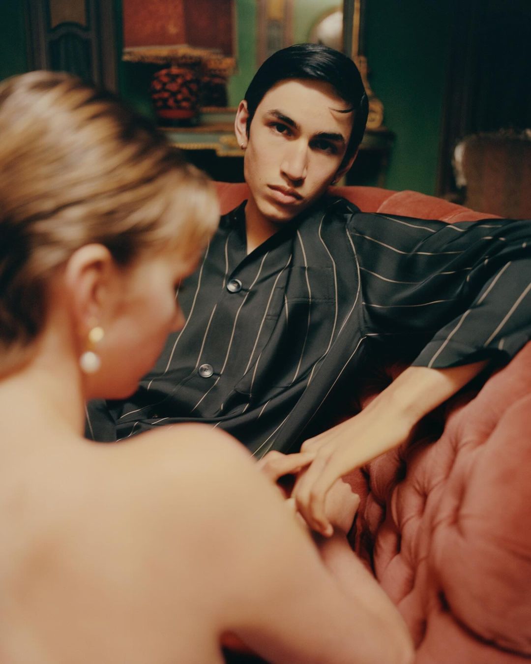 Portrait of a fashionable young man and woman indoors. He is wearing a dark striped shirt. She wears pearl earrings.