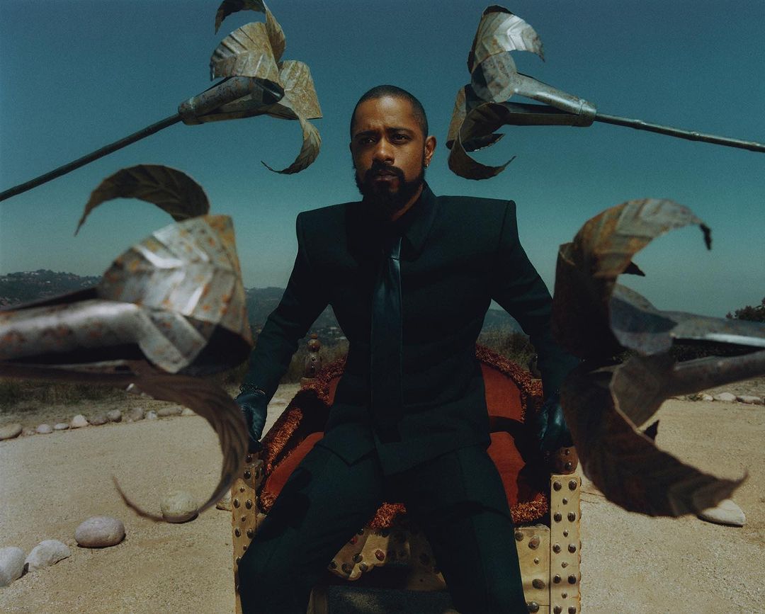 Man in a dark suit sits on an ornate throne outdoors against a desert backdrop with mountains in the distance and blue sky.