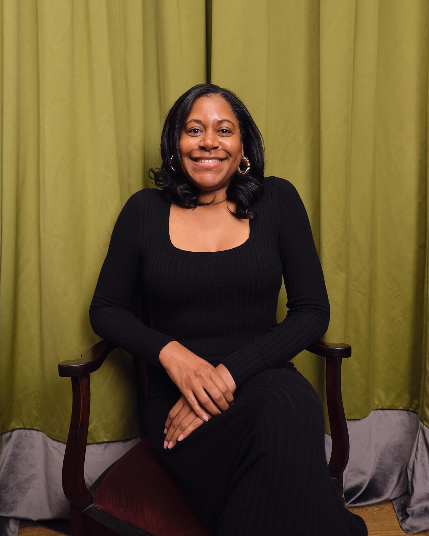 Portrait of a smiling Black woman seated in a wooden armchair against green draped curtains.