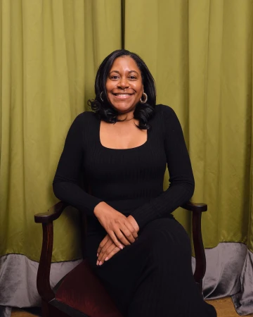 Portrait of a smiling Black woman seated in a wooden armchair against green draped curtains.