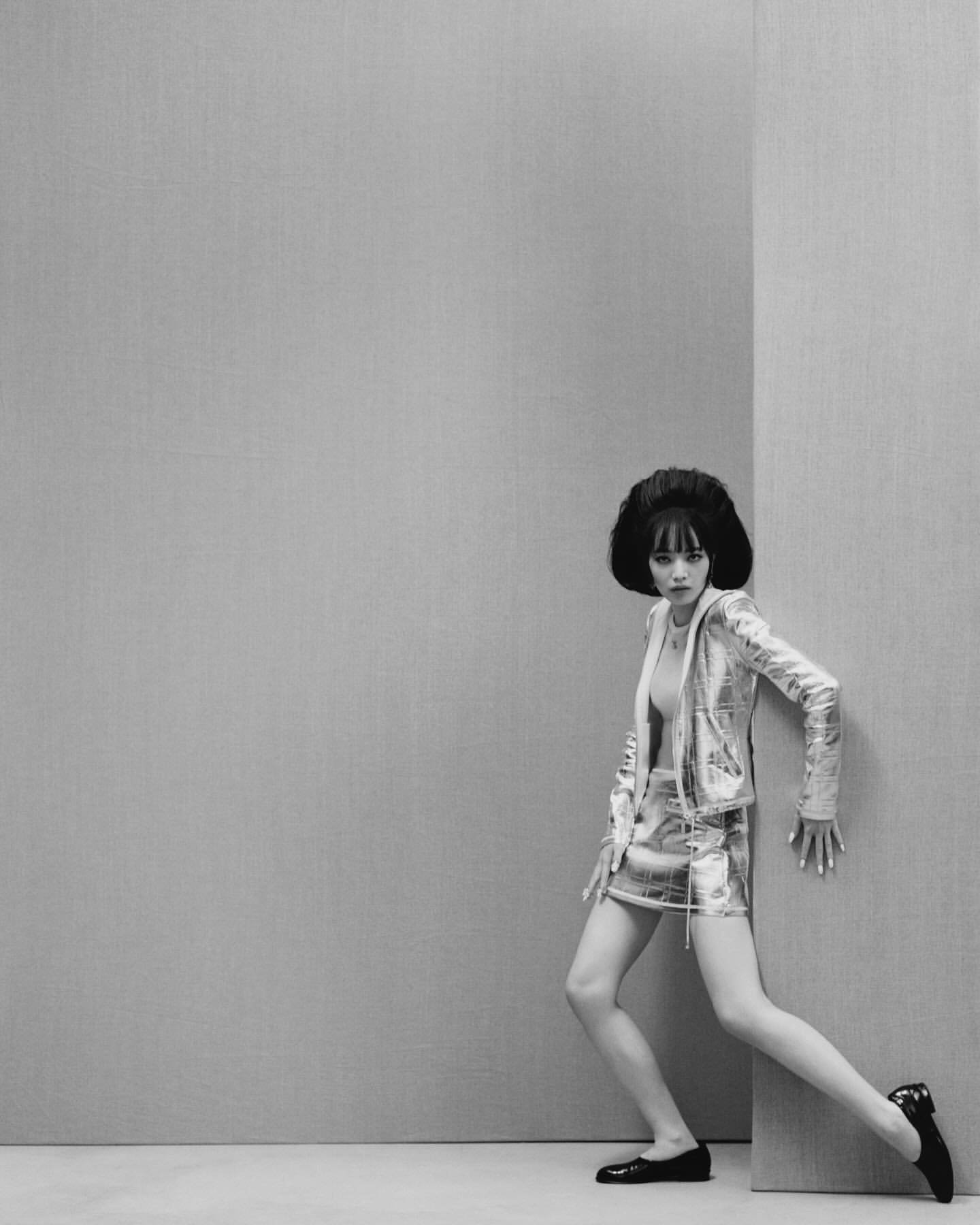 A young woman with a black beehive hairstyle poses in a metallic silver jacket and skirt against a grey wall in a studio setting.