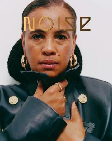 Close-up portrait of a woman wearing a dark leather coat with gold buttons and chunky hoop earrings against a white background.