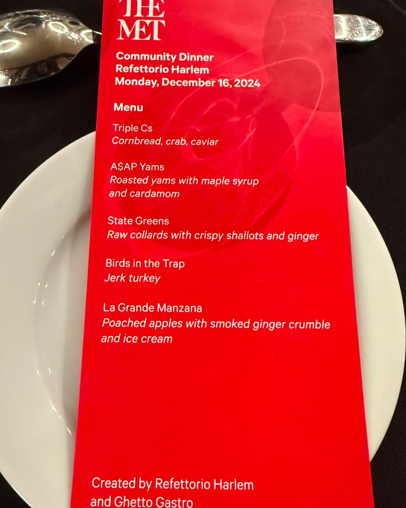Close-up of a bright red menu card listing a multi-course dinner against dark background. Text includes names like Refettorio Harlem.