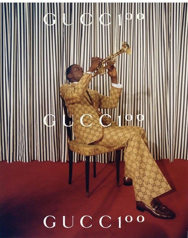 A man plays a trumpet while wearing a Gucci suit, seated in front of a striped backdrop, and the Gucci logo is visible in the scene.